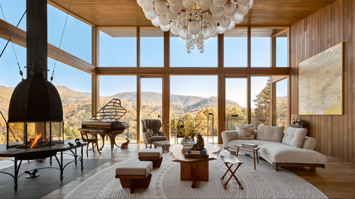 A luxury modern living room with a large white bubble chandelier hanging from a high wood ceiling, featuring a fireplace, grand piano, and a mountain view through floor-to-ceiling windows.