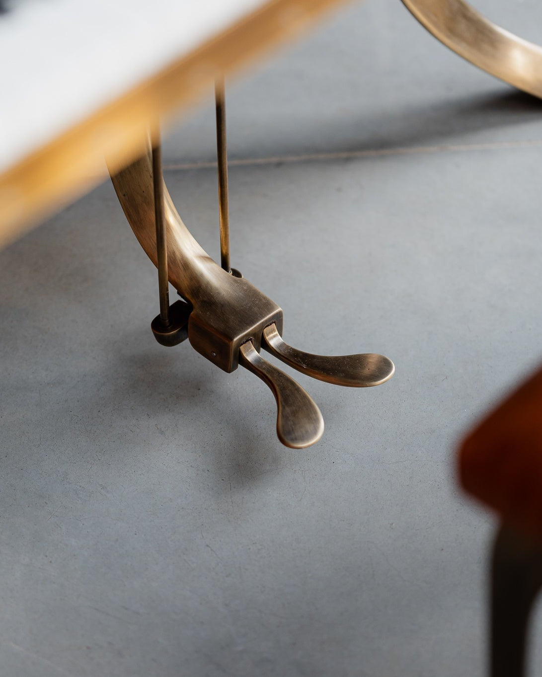 Close-up detail of a brass piano sustain pedal on a gray floor, with parts of a keyboard and a brown leather stool visible in the foreground.