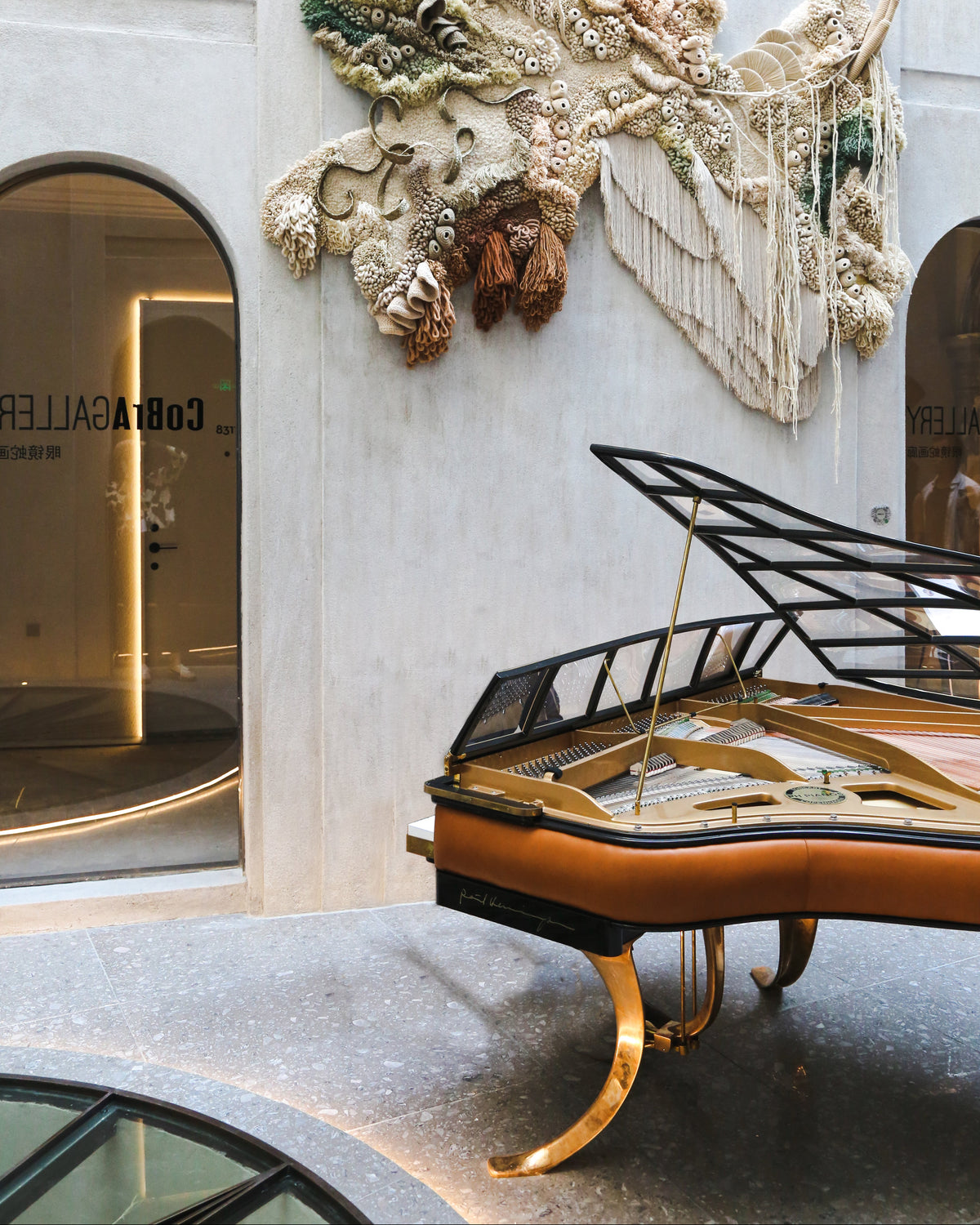A PH grand piano with a tan leather exterior and gold-toned legs is positioned in front of a light gray stone wall featuring a large textured abstract wall installation. A window on the left shows the interior of a gallery space with the text 'COBAGGALLER' visible.